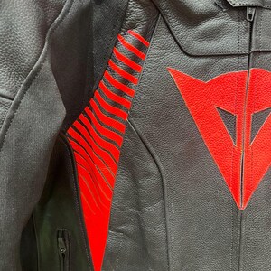 K&ouml;nnte beinhalten: Nahaufnahme einer schwarzen Motorradjacke aus Leder mit roten Akzenten. Die Jacke hat ein rotes Logo auf der Brust und rote Streifen am &Auml;rmel. Sie hat einen Rei&szlig;verschluss und eine kleine Tasche.