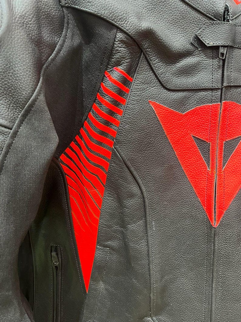 K&ouml;nnte beinhalten: Nahaufnahme einer schwarzen Motorradjacke aus Leder mit roten Akzenten. Die Jacke hat ein rotes Logo auf der Brust und rote Streifen am &Auml;rmel. Sie hat einen Rei&szlig;verschluss und eine kleine Tasche.