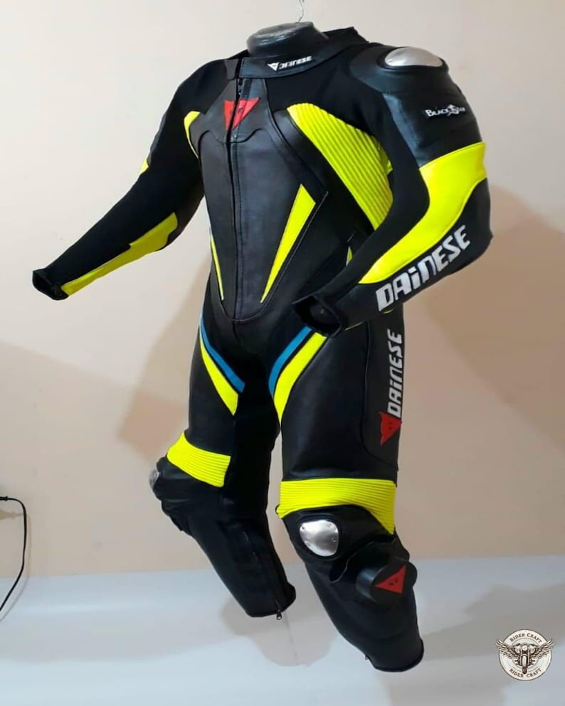K&ouml;nnte beinhalten: Schwarzer und gelber Motorrad-Rennanzug mit dem Markennamen "DAINESSE" auf dem Arm. Der Anzug hat einen Rei&szlig;verschluss vorne, mit roten und blauen Akzenten. Silberne Schutzpolster befinden sich an Schultern und Knien.