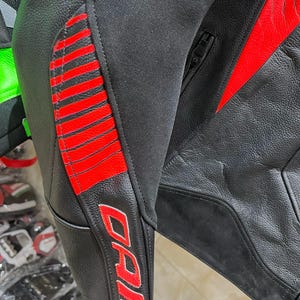 K&ouml;nnte beinhalten: Nahaufnahme einer schwarzen Motorradjacke aus Leder mit roten Akzenten. Die Jacke hat rote Streifen am Arm und den Markennamen "DAI" in roter Schrift. Die Jacke hat einen Rei&szlig;verschluss und besteht aus Leder und Stoff.