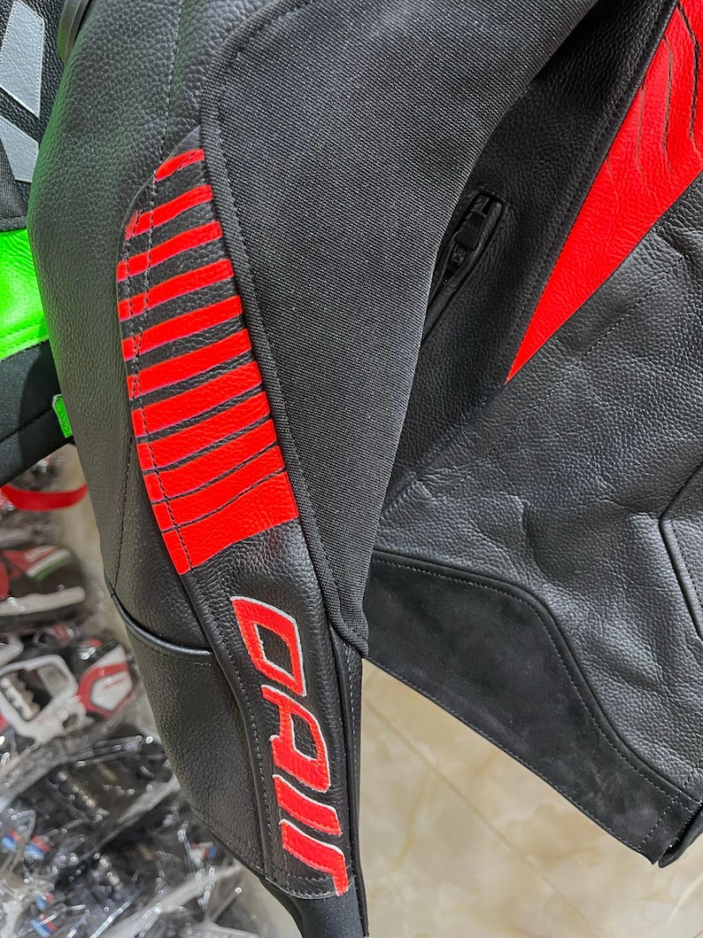 K&ouml;nnte beinhalten: Nahaufnahme einer schwarzen Motorradjacke aus Leder mit roten Akzenten. Die Jacke hat rote Streifen am Arm und den Markennamen "DAI" in roter Schrift. Die Jacke hat einen Rei&szlig;verschluss und besteht aus Leder und Stoff.