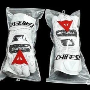 Op de afbeelding: Twee witte motorhandschoenen in een doorzichtige plastic verpakking. Elke handschoen heeft de merknaam "DAINESE" in zwart, een rood driehoeklogo en zwarte accenten. De handschoenen zijn ontworpen voor bescherming en prestaties.
