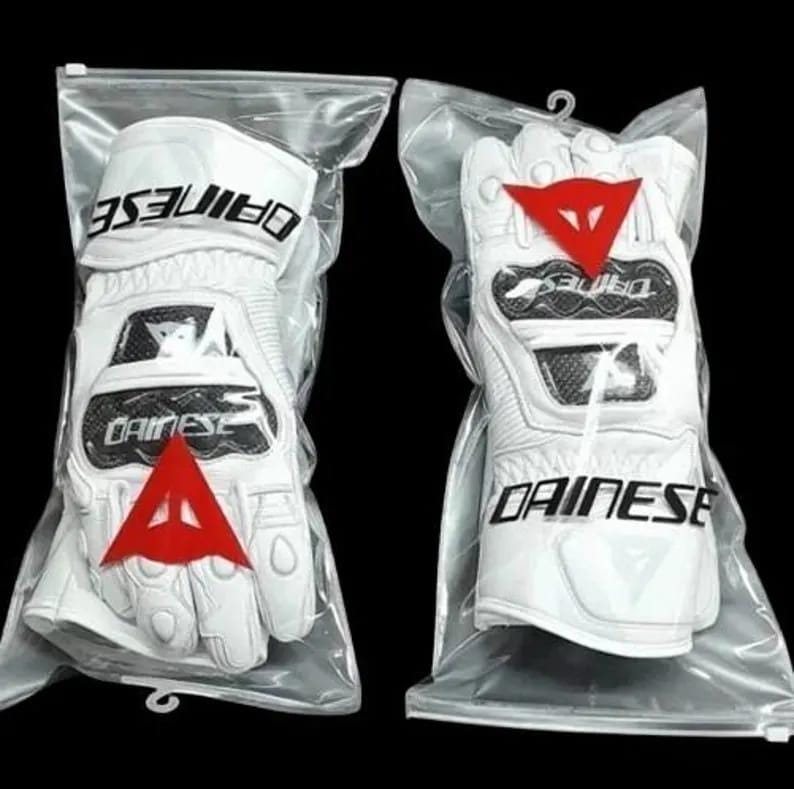 Op de afbeelding: Twee witte motorhandschoenen in een doorzichtige plastic verpakking. Elke handschoen heeft de merknaam "DAINESE" in zwart, een rood driehoeklogo en zwarte accenten. De handschoenen zijn ontworpen voor bescherming en prestaties.