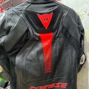 K&ouml;nnte beinhalten: Schwarze Motorradjacke aus Leder mit roten Akzenten. Sie hat ein rotes Dreieckslogo und den Markennamen "DAINESE" in roter Schrift. Gerippte Details sind auf den Schultern und dem R&uuml;cken zu sehen.