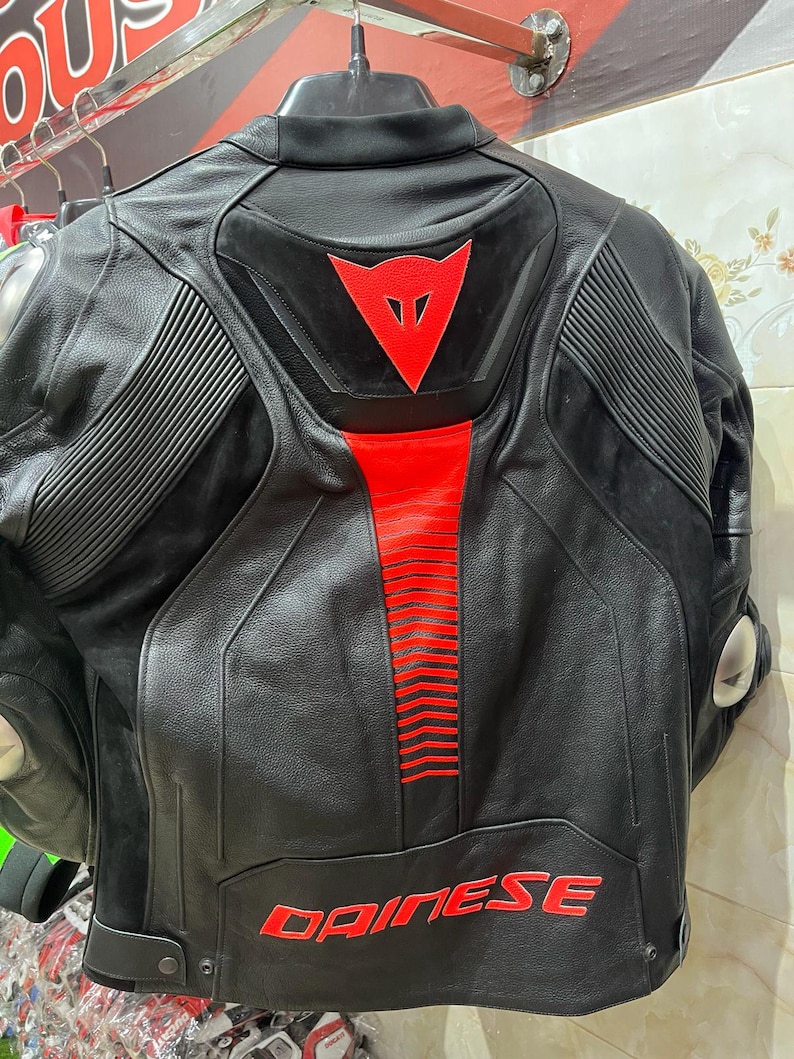 K&ouml;nnte beinhalten: Schwarze Motorradjacke aus Leder mit roten Akzenten. Sie hat ein rotes Dreieckslogo und den Markennamen "DAINESE" in roter Schrift. Gerippte Details sind auf den Schultern und dem R&uuml;cken zu sehen.