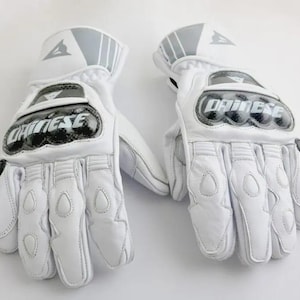 Op de afbeelding: Paar witte motorhandschoenen met de merknaam "Dainese" op de knokkels. De handschoenen hebben verstevigde padding op de handpalmen en vingers, met grijze accenten op de manchetten. Ontworpen voor bescherming en prestaties.