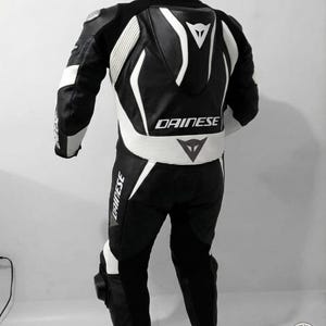 K&ouml;nnte beinhalten: Schwarz-wei&szlig;er Motorrad-Rennanzug mit dem Markennamen "DAINESE" auf dem R&uuml;cken, der Taille und dem Bein. Der Anzug hat wei&szlig;e Akzente und Polsterungen an Schultern, Armen und Beinen. Der Anzug h&auml;ngt an einem Kleiderb&uuml;gel.