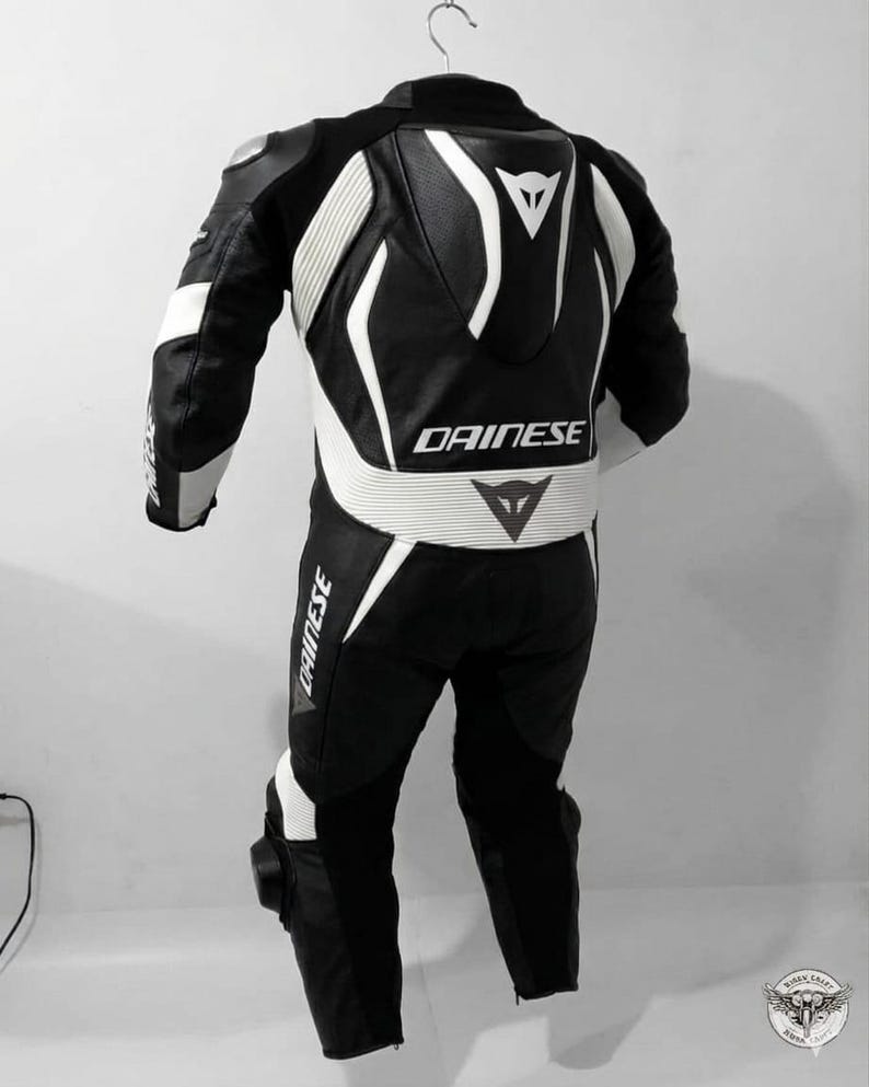 K&ouml;nnte beinhalten: Schwarz-wei&szlig;er Motorrad-Rennanzug mit dem Markennamen "DAINESE" auf dem R&uuml;cken, der Taille und dem Bein. Der Anzug hat wei&szlig;e Akzente und Polsterungen an Schultern, Armen und Beinen. Der Anzug h&auml;ngt an einem Kleiderb&uuml;gel.