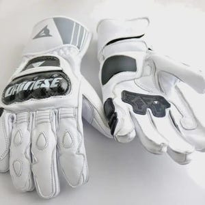 Op de afbeelding: Een paar witte motorhandschoenen met zwarte en grijze accenten. De handschoenen hebben beschermende padding op de knokkels en handpalmen, en het merk "Dainese" is prominent aanwezig.