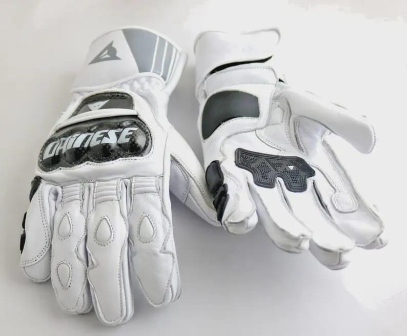 Op de afbeelding: Een paar witte motorhandschoenen met zwarte en grijze accenten. De handschoenen hebben beschermende padding op de knokkels en handpalmen, en het merk "Dainese" is prominent aanwezig.