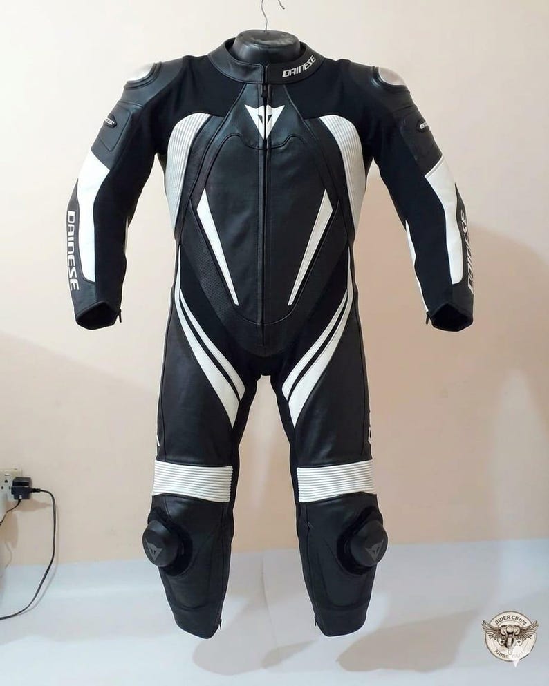 K&ouml;nnte beinhalten: Schwarzer und wei&szlig;er Motorrad-Rennanzug aus Leder mit dem Markennamen "Dainese" auf Brust und Armen. Der Anzug hat wei&szlig;e Streifen und Polsterungen an Schultern, Armen und Beinen. Knieschleifer sind sichtbar.