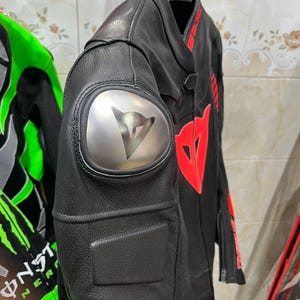 K&ouml;nnte beinhalten: Schwarze Motorradjacke aus Leder mit roten Akzenten und einem silbernen Schulterpolster mit Logo. Die Jacke hat ein stromlinienf&ouml;rmiges Design mit gepolsterten Bereichen und h&auml;ngt an einem schwarzen Kleiderb&uuml;gel.