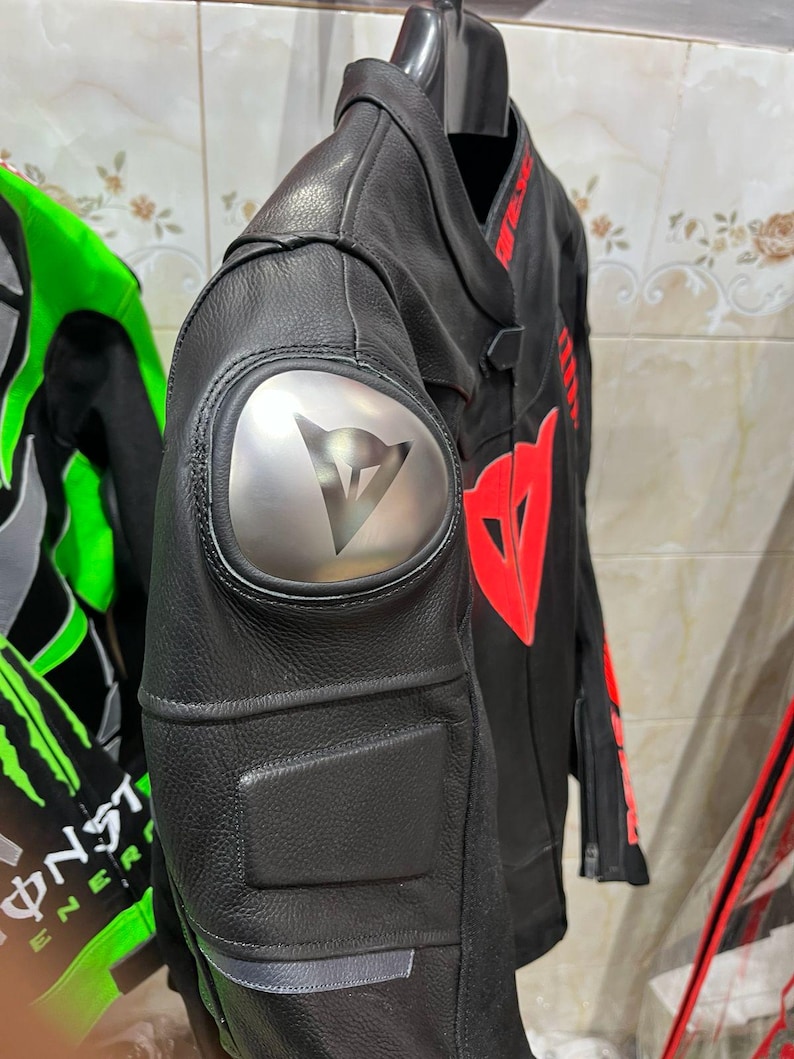 K&ouml;nnte beinhalten: Schwarze Motorradjacke aus Leder mit roten Akzenten und einem silbernen Schulterpolster mit Logo. Die Jacke hat ein stromlinienf&ouml;rmiges Design mit gepolsterten Bereichen und h&auml;ngt an einem schwarzen Kleiderb&uuml;gel.