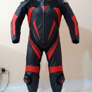 K&ouml;nnte beinhalten: Schwarzer und roter Motorrad-Rennanzug mit dem Markennamen "Dainese". Der Anzug hat rote Akzente an Armen, Beinen und Torso sowie Protektoren an Schultern und Knien.