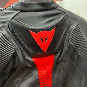 K&ouml;nnte beinhalten: Schwarze Motorradjacke aus Leder mit roten Akzenten. Die Jacke hat ein rotes Dreieckslogo auf dem R&uuml;cken und rote Streifen entlang der Wirbels&auml;ule. Sie hat gerippte Eins&auml;tze an den Schultern und einen gepolsterten R&uuml;cken.