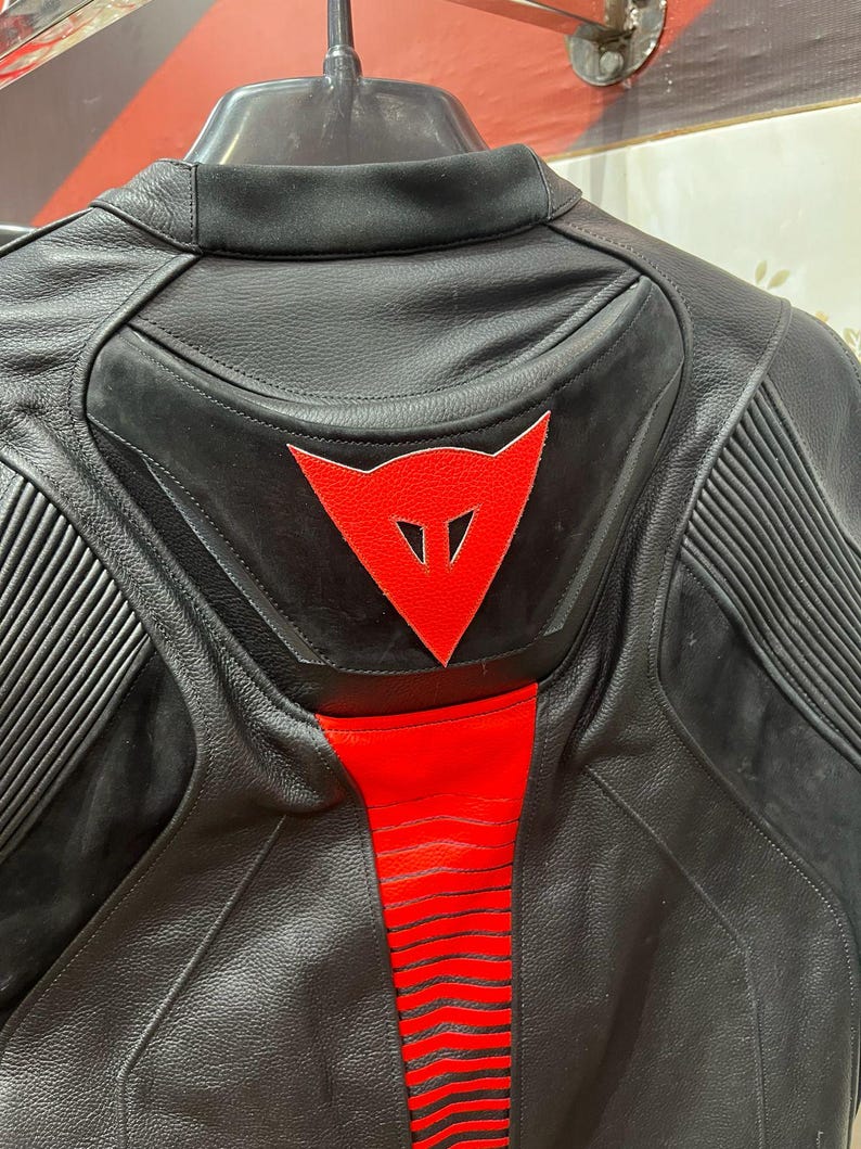 K&ouml;nnte beinhalten: Schwarze Motorradjacke aus Leder mit roten Akzenten. Die Jacke hat ein rotes Dreieckslogo auf dem R&uuml;cken und rote Streifen entlang der Wirbels&auml;ule. Sie hat gerippte Eins&auml;tze an den Schultern und einen gepolsterten R&uuml;cken.