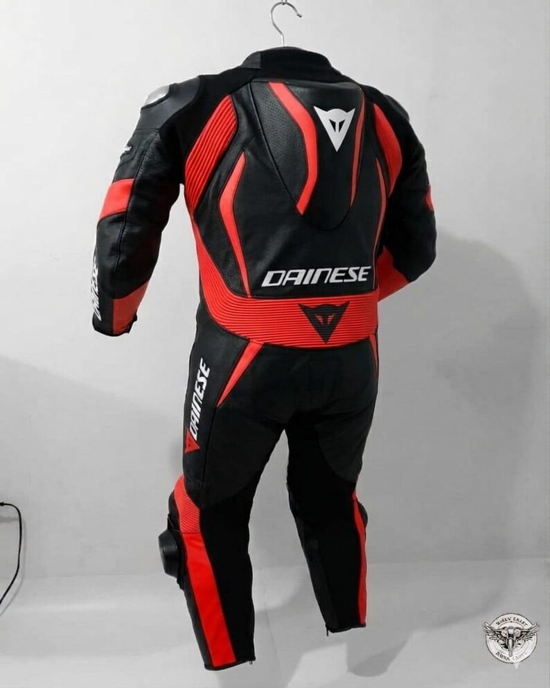 K&ouml;nnte beinhalten: Schwarzer und roter Motorrad-Rennanzug mit dem Markennamen "DAINESE". Der Anzug hat ein stromlinienf&ouml;rmiges Design mit roten Akzenten an Schultern, Armen und Beinen. Der Anzug h&auml;ngt an einem Kleiderb&uuml;gel.
