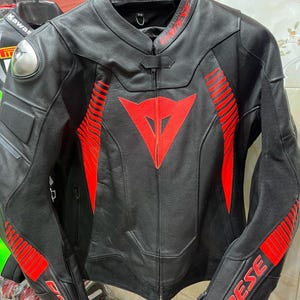 K&ouml;nnte beinhalten: Schwarze Motorradjacke aus Leder mit roten Akzenten. Sie hat ein rotes Dreieckslogo auf der Brust und rote Streifen auf den &Auml;rmeln. Die Jacke hat einen Rei&szlig;verschluss und einen Stehkragen. Der Markenname ist sichtbar.