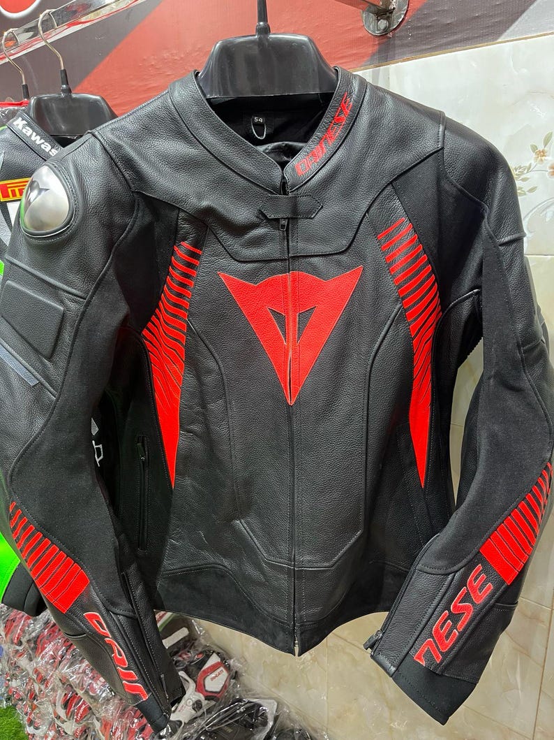 K&ouml;nnte beinhalten: Schwarze Motorradjacke aus Leder mit roten Akzenten. Sie hat ein rotes Dreieckslogo auf der Brust und rote Streifen auf den &Auml;rmeln. Die Jacke hat einen Rei&szlig;verschluss und einen Stehkragen. Der Markenname ist sichtbar.