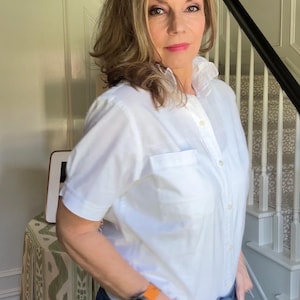 May include: A crisp white short-sleeved button-down shirt featuring a classic collar, a single chest pocket, and a front button placket. The shirt is paired with dark blue denim jeans and a digital watch with a bright orange silicone band.