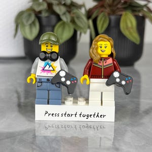 Puede incluir: Dos minifiguras de plástico sobre una base rectangular blanca con el texto Press start together. La figura de la izquierda lleva una gorra verde, camiseta blanca con un triángulo colorido y pantalones azules. La derecha tiene pelo ondulado y chaqueta roja.