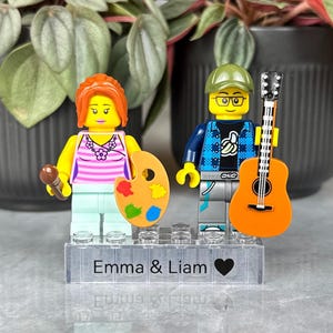 Könnte beinhalten: Zwei individuelle LEGO-Minifiguren auf einem transparenten rechteckigen Sockel mit der Aufschrift Emma & Liam und einem Herzsymbol. Die linke Figur hat orangefarbenes Haar, ein rosa gestreiftes Oberteil und hält einen Pinsel und eine Palette. Die rechte Figur trägt eine grüne Kappe, eine Brille, ein blaues Karohemd und hält eine orangefarbene Akustikgitarre.