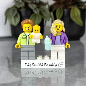 Może przedstawiać: Dwie minifigurki Lego stojące na białej prostokątnej podstawie z napisem The Smith Family i ikoną serca. Jedna figurka ma brązowe włosy, jasnozieloną kurtkę i szare spodnie, trzymając małe żółte niemowlę. Druga ma blond włosy, fioletowy kardigan i białe spodnie, trzymając jasnoniebieską butelkę dla niemowląt.