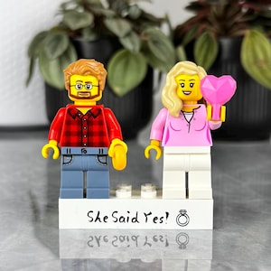 Puede incluir: Dos minifiguras de plástico sobre una base rectangular blanca. La figura de la izquierda tiene pelo castaño, barba, gafas, camisa de cuadros roja y pantalones azules. La de la derecha tiene pelo rubio, camisa rosa y pantalones blancos, sosteniendo un corazón rosa.