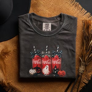 May include: A charcoal gray short-sleeve t-shirt featuring a graphic of three red Coca-Cola cans with black and white patterned bows and straws. The design includes a white ghost, two orange jack-o'-lanterns, and a black haunted house silhouette.