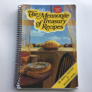 May include: A spiral-bound cookbook titled The Mennonite Treasury of Recipes with a cover illustration of a baked pie on a windowsill, a table with a plate, a mug, and a napkin, and a red Canadian Best Seller seal in the upper right corner.