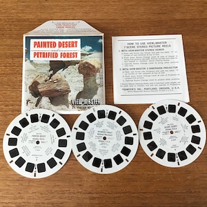 May include: A View-Master set featuring a cardboard envelope titled Painted Desert and Petrified Forest, an instructional paper, and three white circular reels with black film frames. Each reel contains seven stereo images of Arizona landscapes.