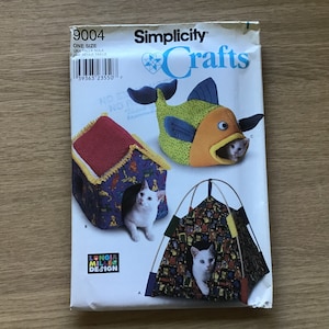 May include: Simplicity Crafts pattern 9004 featuring three cat bed designs: a house, a fish, and a tent. The pattern includes a house with a red roof, a fish with orange and green details, and a tent with a colorful print. A white cat is visible in each bed.
