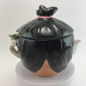 May include: A black and brown ceramic teapot shaped like a person with a black hair bun and a pink ribbon. The teapot has a white spout and a green and white handle.