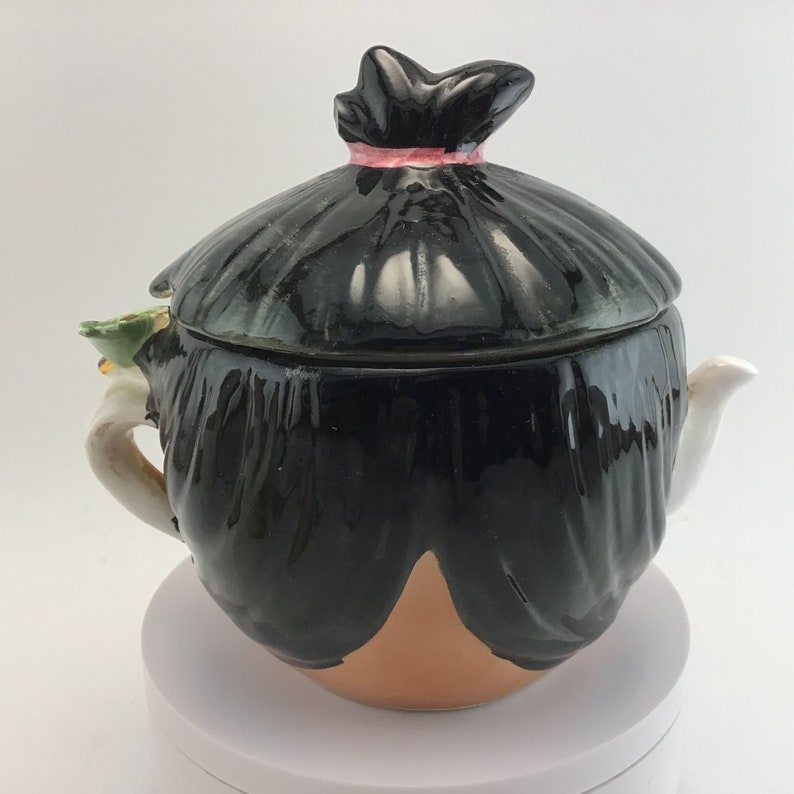 May include: A black and brown ceramic teapot shaped like a person with a black hair bun and a pink ribbon. The teapot has a white spout and a green and white handle.