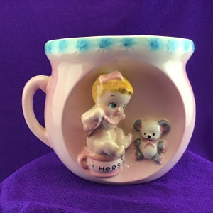 May include: A pink ceramic baby cup with a handle, featuring a sculpted baby figure and a teddy bear. The cup has a light blue floral design around the rim and the word "HERS" on the potty. A charming vintage baby item.