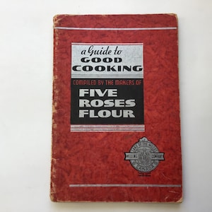 May include: A vintage cookbook titled "a Guide to Good Cooking" with a red cover. The cover features the text "Compiled by the Makers of Five Roses Flour" in a black and white design. The book also has a logo for Lake of the Woods Milling Company.