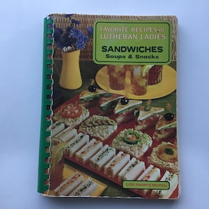May include: A vintage cookbook titled "Favorite Recipes of Lutheran Ladies" with a green spiral binding. The cover features a green rectangle with the title "Sandwiches, Soups & Snacks." The image shows various sandwiches and a yellow vase with blue flowers.