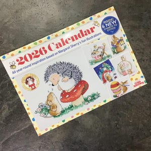 以下が含まれることがあります： マーガレット・シェリーのイラストが特徴の2026年カレンダー。表紙にはハリネズミと赤いキノコを持つネズミが描かれています。カレンダーには、6つの新しいクロスステッチチャートが含まれています。ボーダーは、カラフルな水玉模様の黄色です。