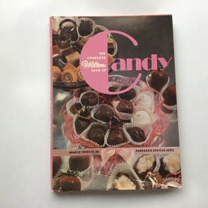 May include: A vintage cookbook titled "The Complete Wilton Book of Candy" with a pink and black cover. The cover features a pink circle with the word "Candy" and images of chocolates and candies.