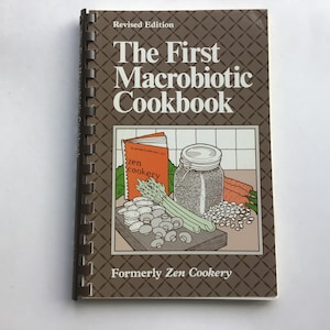 May include: A spiral-bound cookbook titled The First Macrobiotic Cookbook, formerly Zen Cookery. The brown cover features a diamond pattern and an illustration of a kitchen scene with a jar, mushrooms, asparagus, carrots, and a book.
