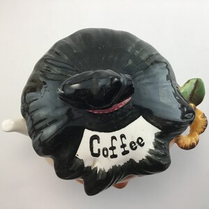 May include: A ceramic coffee pot shaped like a woman wearing a black hat and a white apron with the word "Coffee" written on it.
