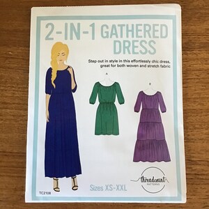 May include: A sewing pattern for a 2-in-1 gathered dress. The pattern features illustrations of the dress in three different styles, including a long blue dress, a green dress, and a purple dress. The text on the pattern reads "2-IN-1 GATHERED DRESS."