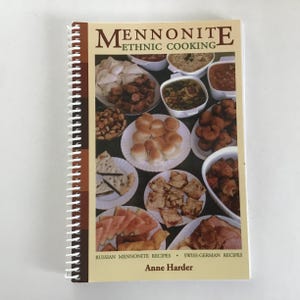 May include: A spiral-bound cookbook titled "Mennonite Ethnic Cooking" with a cream-colored cover. The cover features a collage of various dishes. The book includes recipes from Russian Mennonite and Swiss-German traditions, authored by Anne Harder.