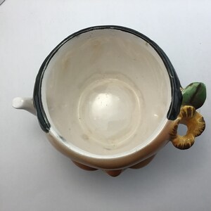 May include: A white ceramic teapot with a black rim and a brown, flower-shaped handle. The teapot has a green leaf and a yellow flower on the handle.