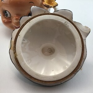 May include: The inside of a ceramic cookie jar lid with a brown cork lining and a white glaze. The bottom of the lid has a small, white, circular mark with a letter "G" in the center.