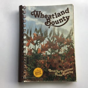 Peut inclure: Un livre de cuisine à reliure spirale intitulé Wheatland Bounty avec une couverture illustrée de lys orange et de tiges de blé sous un ciel nuageux. La couverture porte le texte Good Old Fashioned Home Cooking et une étiquette étoilée jaune.