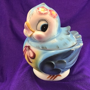 May include: A blue and yellow ceramic bird figurine with a white base. The bird has a pink and yellow flower on its head and a red ribbon around its neck. The bird is a sugar bowl with a lid.
