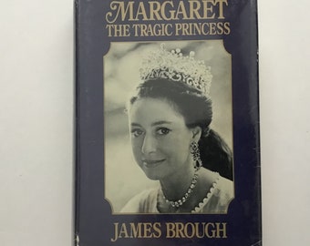 Może przedstawiać: Książka w twardej oprawie zatytułowana Margaret The Tragic Princess autorstwa Jamesa Brougha. Ciemnoniebieska okładka zawiera czarno-biały portret kobiety w wysadzanej klejnotami tiarze, kolczykach i naszyjniku, ujęty w złotą ramkę.