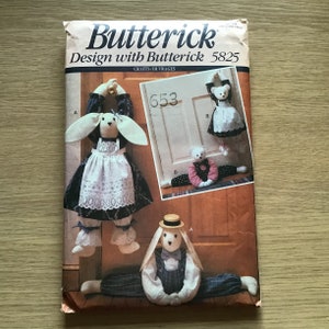 May include: A Butterick 5825 sewing pattern envelope featuring illustrations of fabric dolls. One doll is a rabbit wearing a dark dress and white apron, another is a bear in a dress, and a third is a rabbit in a hat and overalls.
