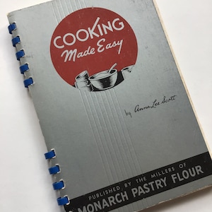 May include: A vintage cookbook, "COOKING Made Easy" by Anna Lee Scott. The cover is grey with a red circle featuring the title in white. The book is bound with blue rings and published by Monarch Pastry Flour.